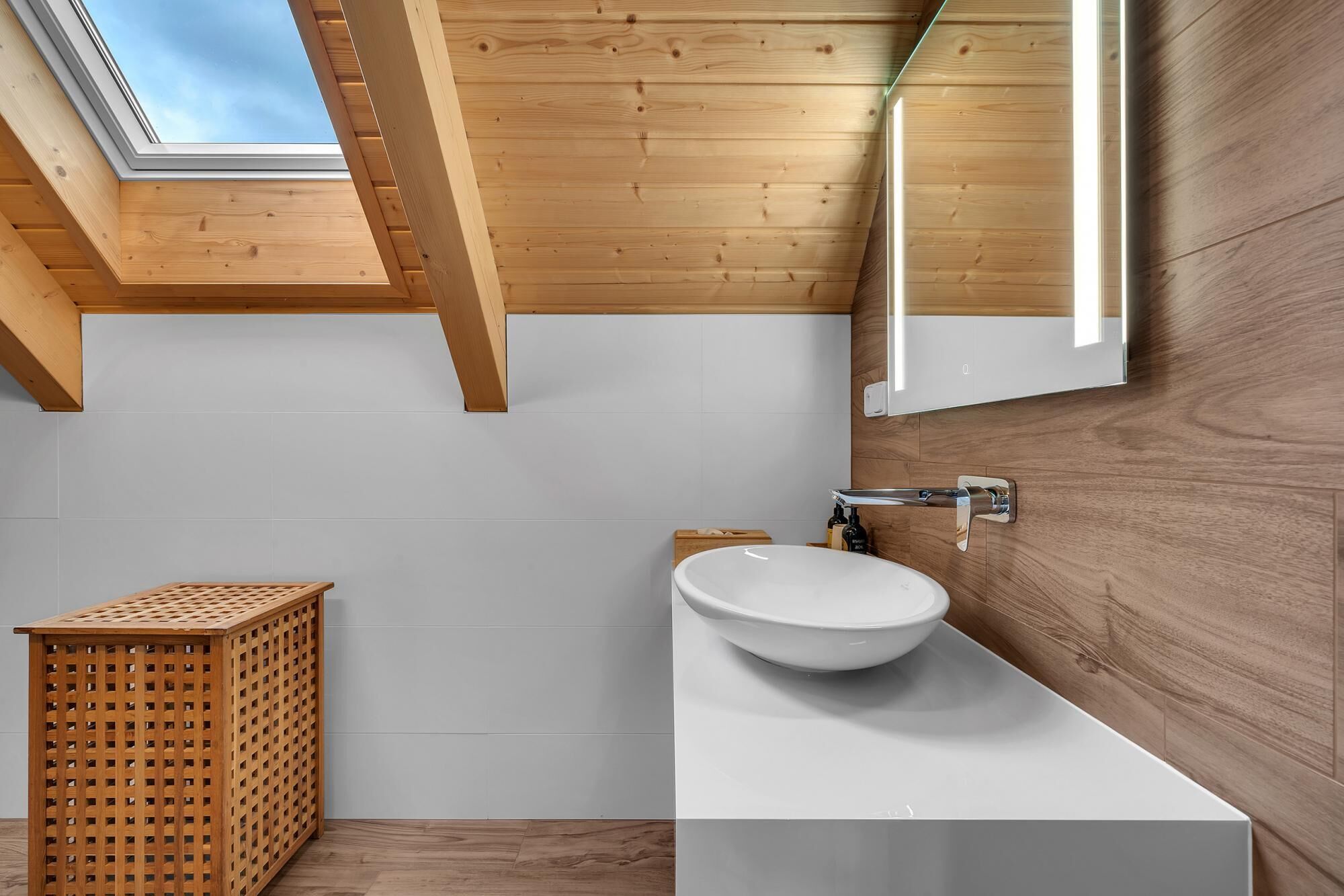 A modern bathroom with a skylight, wooden ceiling, white tiles, a large mirror, and a wooden laundry basket.
