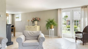 A spacious living room with a light-colored sofa, a small white side table with a lamp and flowers, a potted plant, and a view through a glass door to the outside. - Cozy cottage with heated pool, garden and covered terrace (Tzummarum)
