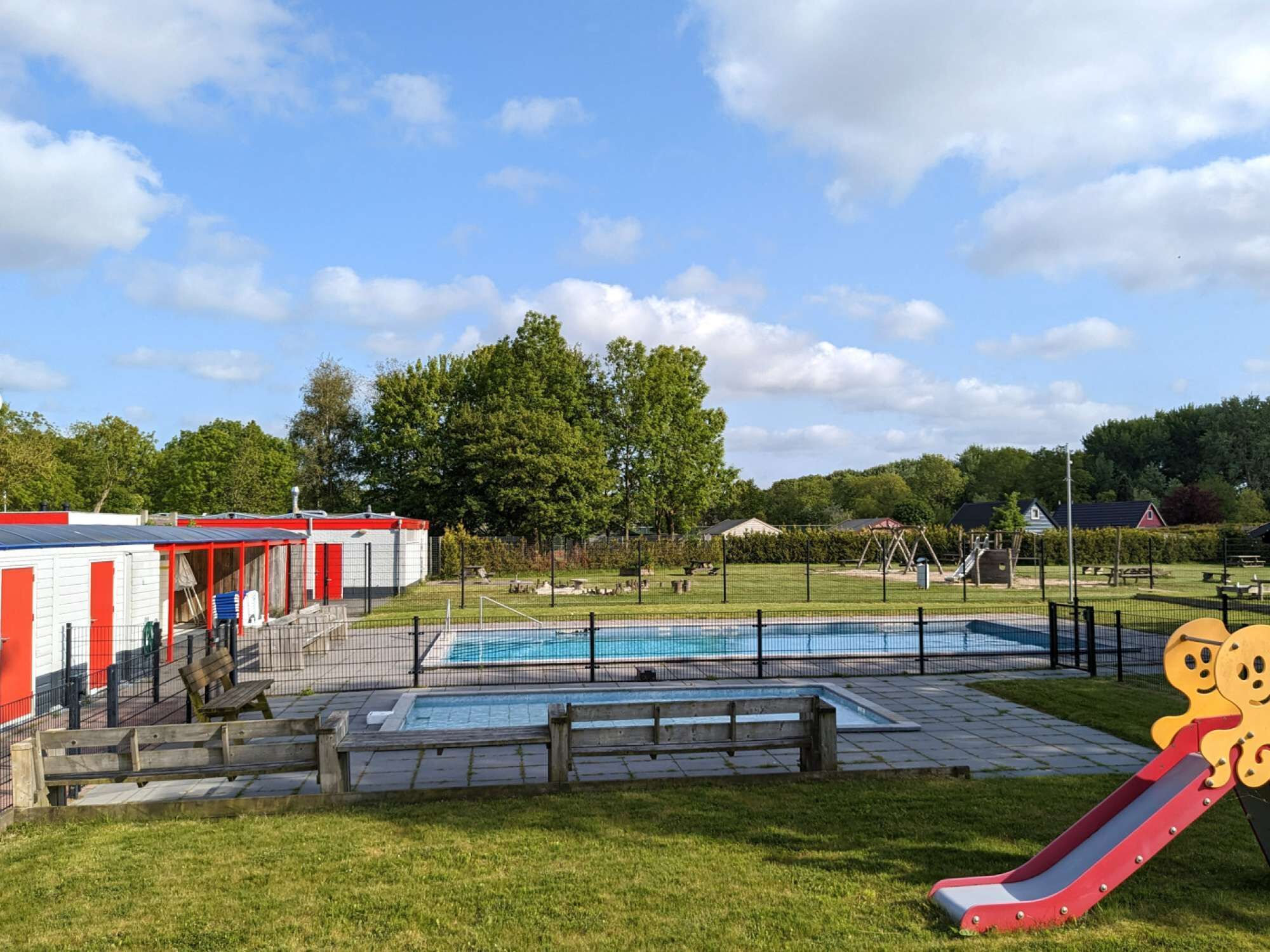 Ein Schwimmbeckenbereich mit Rutsche, umgeben von einem eingezäunten Spielplatz und einem Gebäude mit roter und weißer Fassade.