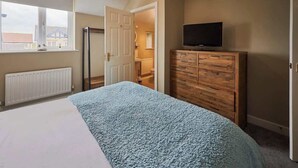 Ein Schlafzimmer mit einem Bett, einer Holzkommode, einem Fernseher darauf und einem Fenster mit Blick auf Gebäude.