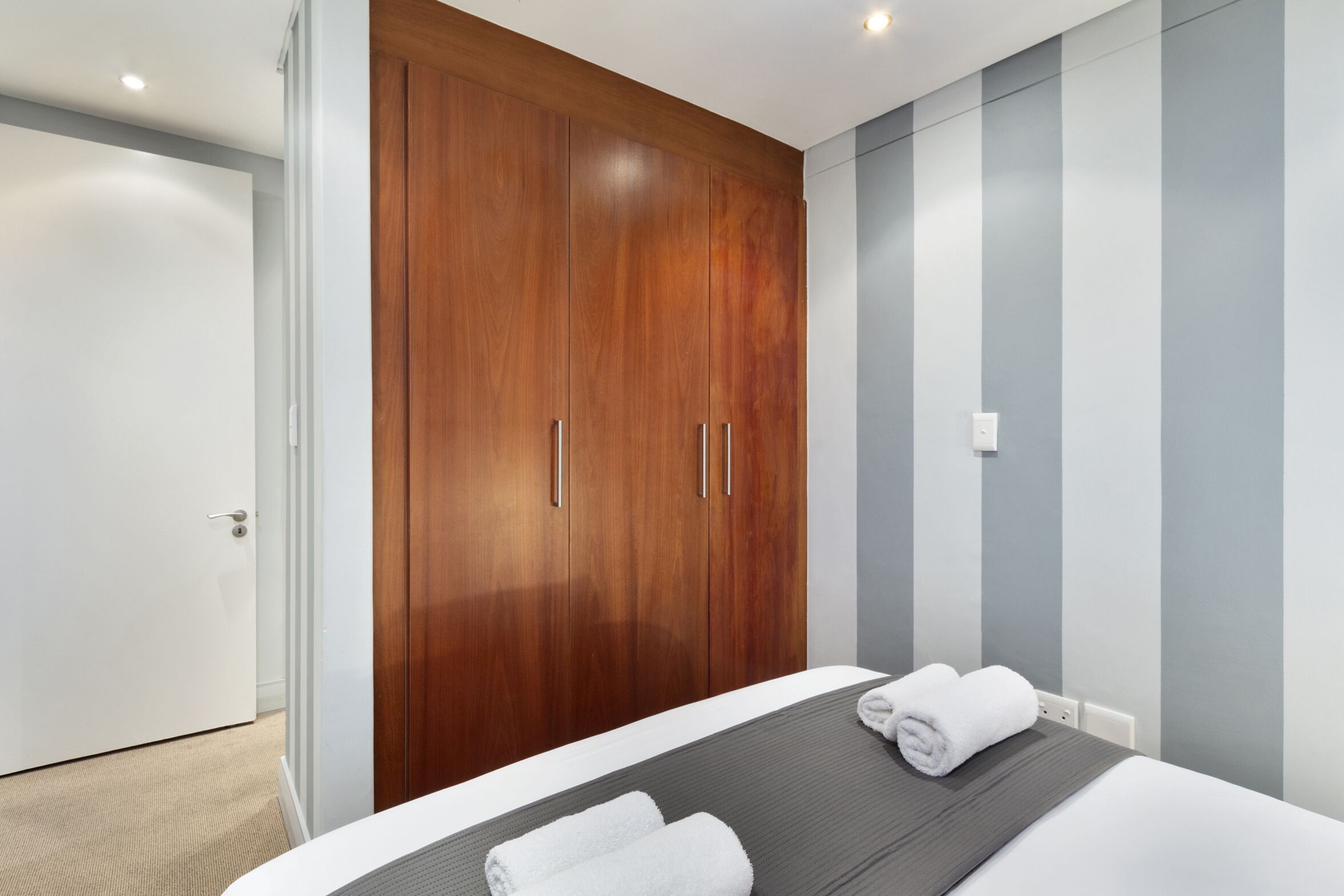 Une chambre d’hôtel moderne, dotée d’une grande armoire en bois, d’un lit soigneusement fait avec du linge de maison blanc et d’un mur à rayures.