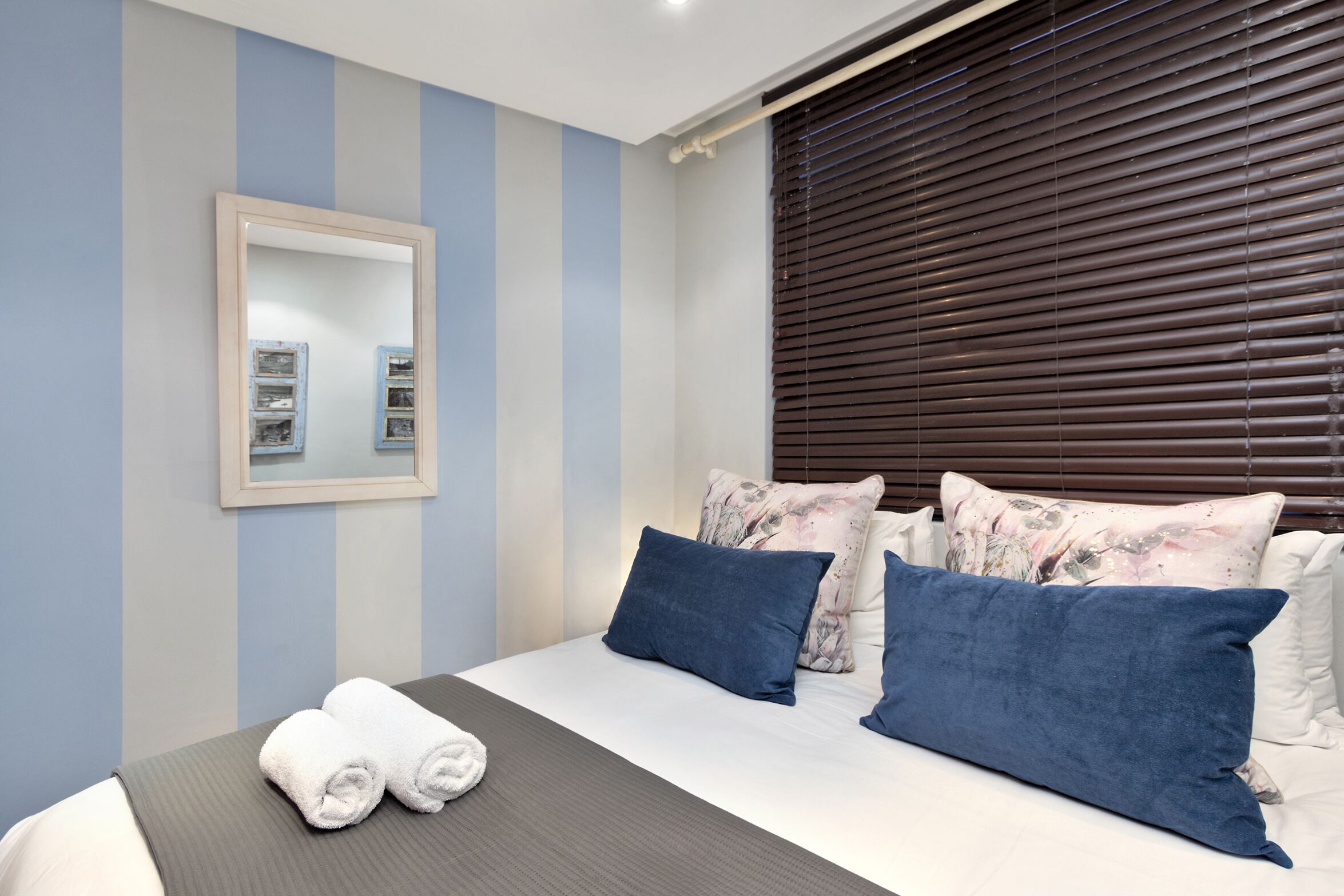 A neatly made bed with white and blue bedding, a mirror on the wall, and a window with blinds.