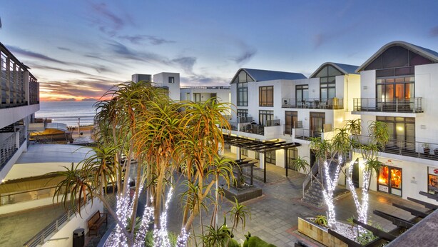 A hotel courtyard with a view of the ocean at dusk, featuring modern architecture, outdoor seating, and palm trees. - Eden on the Bay 129 (Cape Town)