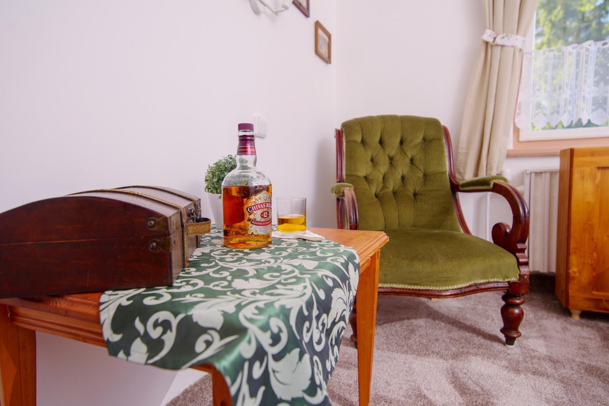 A room with a wooden table, a bottle of Chivas Regal, a glass of whiskey, a green armchair, and a wooden cabinet.