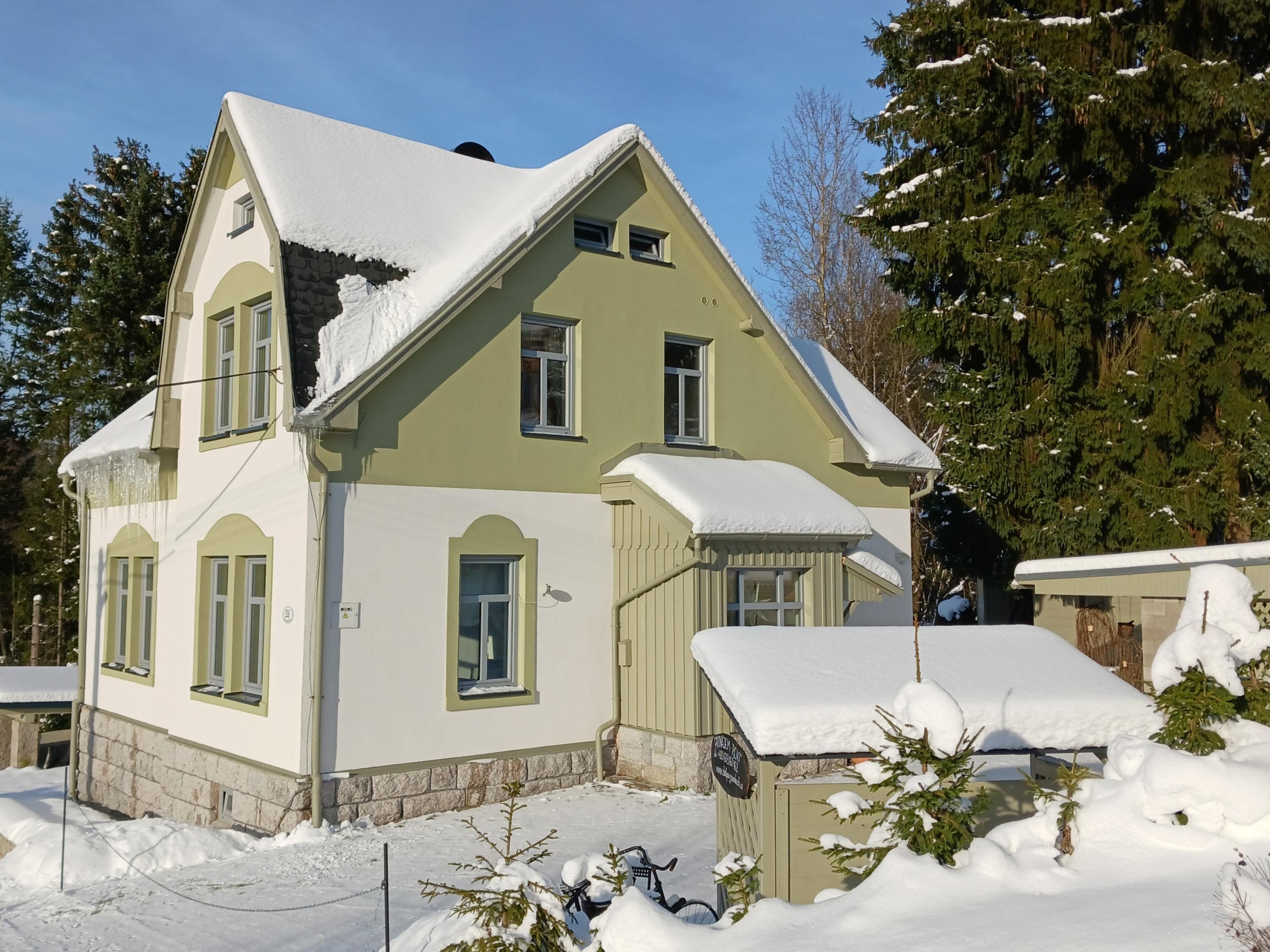 A two-story house with a steeply pitched roof covered in snow, surrounded by snow-covered trees and a clear sky.