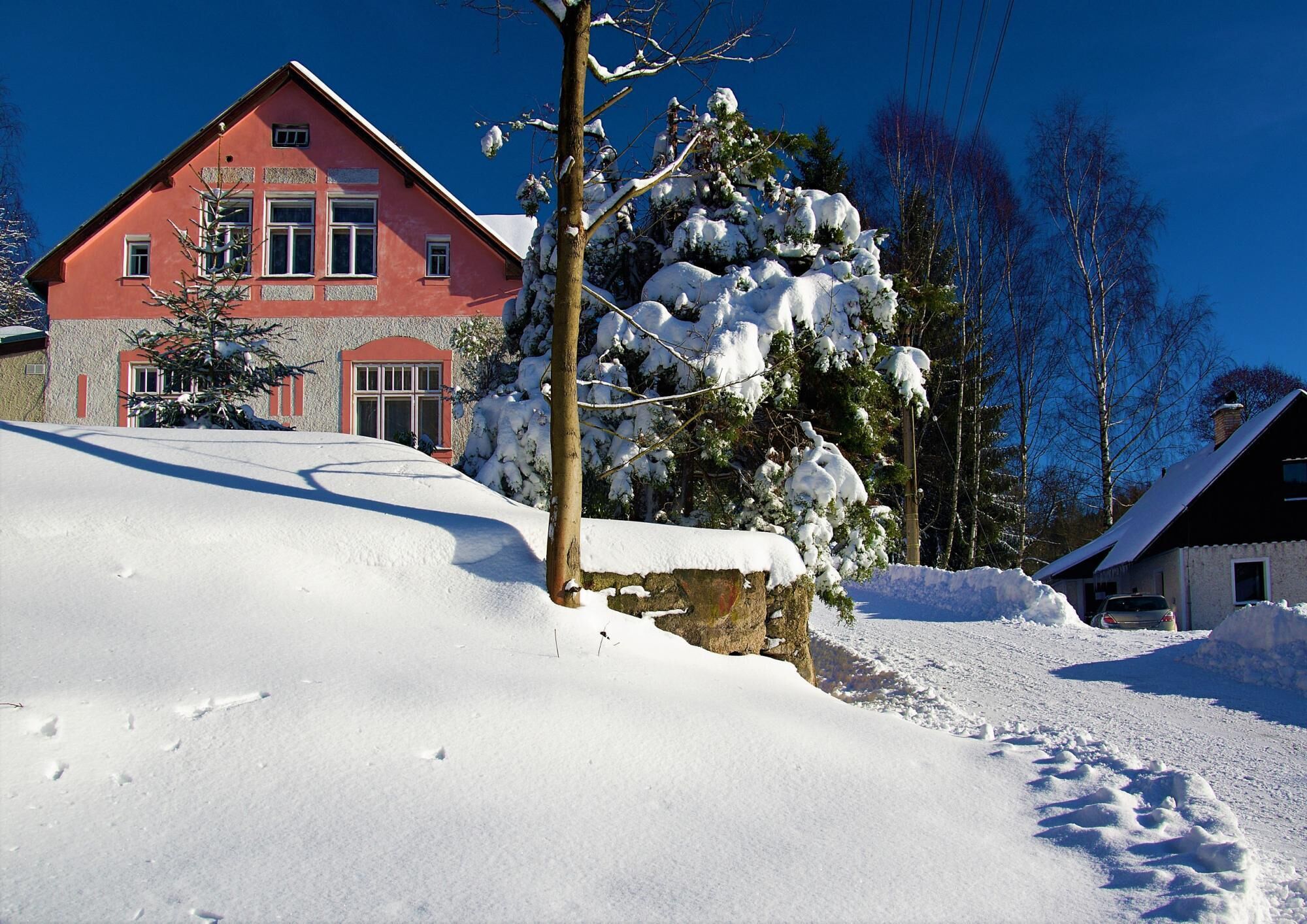 Paesaggio innevato con un edificio rosso, un muro di pietra e un albero piegato dalla neve.