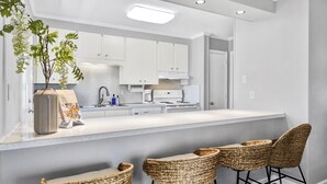Une cuisine moderne avec des meubles blancs, un long comptoir et une cuisinière intégrée. On y trouve trois chaises en osier près du comptoir.