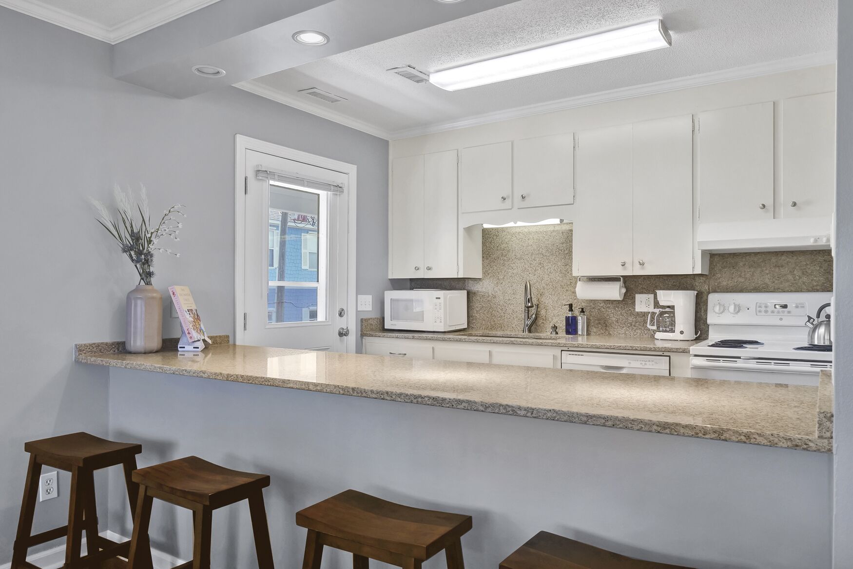 Una cocina moderna con armarios blancos, encimera de granito, microondas empotrado, pileta de acero inoxidable y estufa. Hay tres taburetes de madera en la barra.
