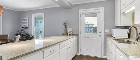 A modern kitchen with white cabinetry, a granite countertop, a built-in microwave, a sink, and a view of the outside through a door.