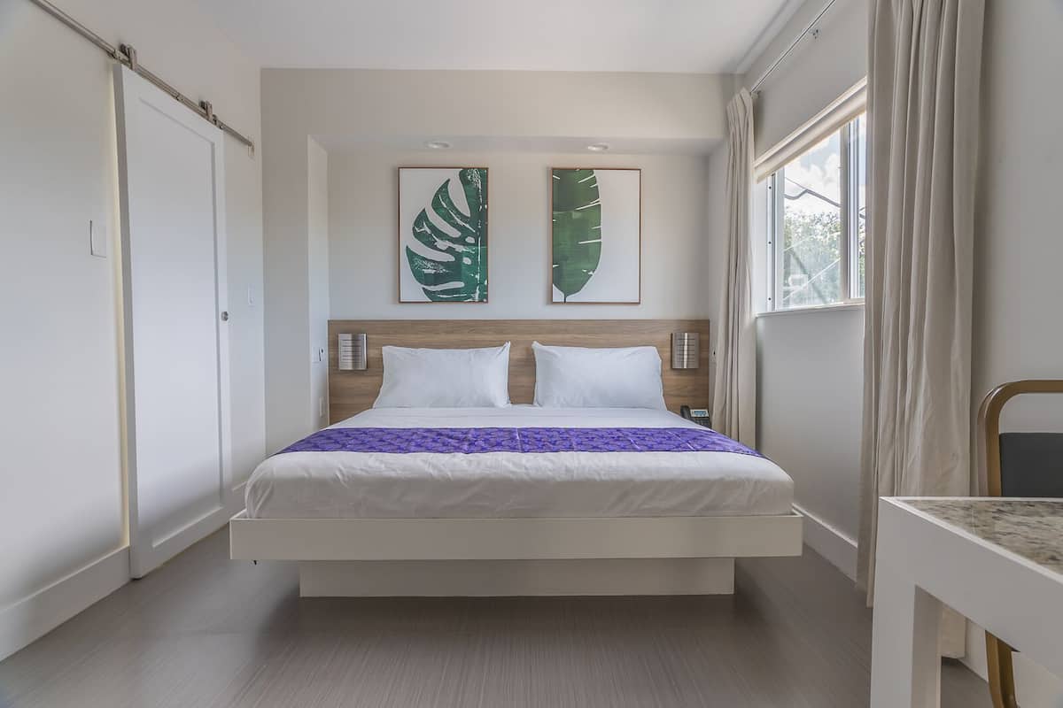 Quarto de hotel moderno com uma cama grande, duas obras de arte penduradas na parede, telefone ao lado da cama e uma janela com cortinas.