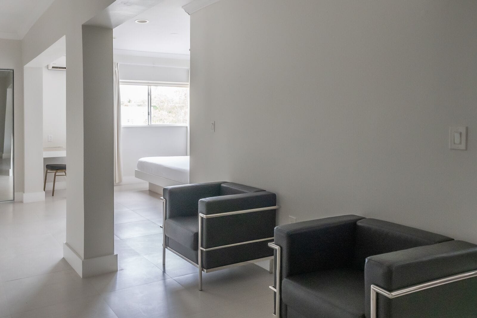 A modern, minimalist room with a large window, a bed, and two black armchairs.