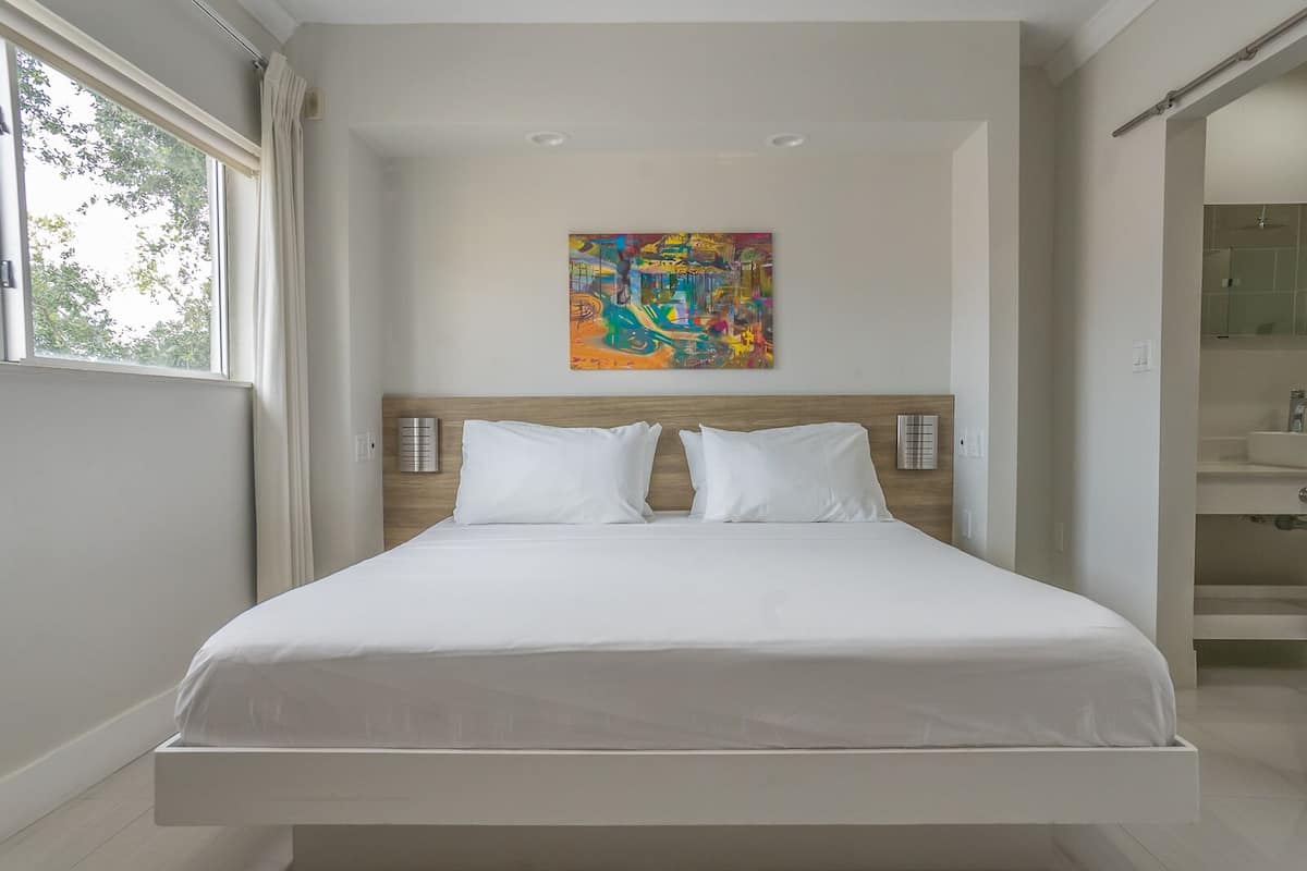 Quarto de hotel moderno com uma cama grande, uma pintura abstrata colorida acima dela e vista para o banheiro por uma porta aberta.