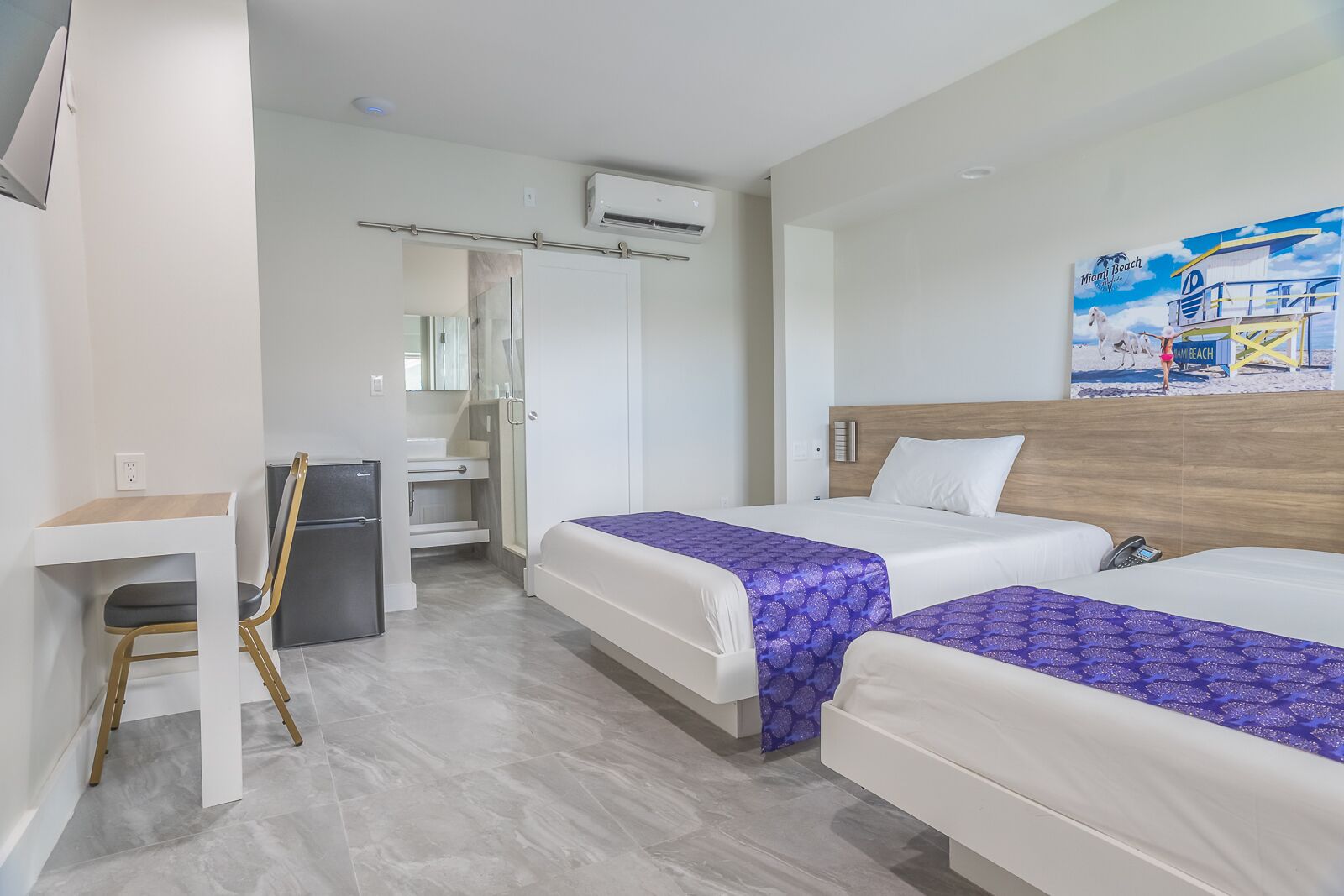 Habitación de hotel con dos camas, un escritorio, una silla, un refrigerador y un baño con ducha e inodoro.