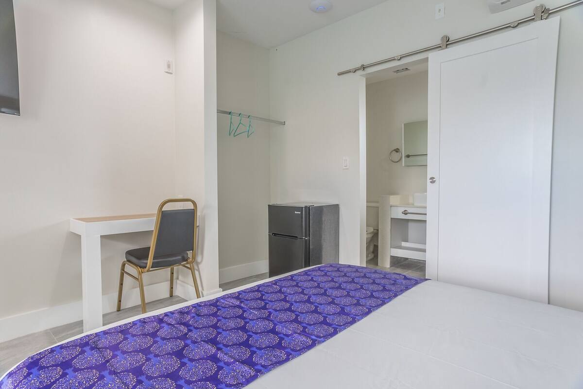 Quarto com cama, escrivaninha, cadeira, frigorífico e banheiro visível por uma porta aberta.