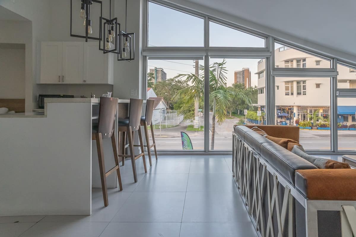 Uma cozinha moderna com armários brancos, um espaço de bar com banquetas e uma janela grande com vista para a rua e edifícios.