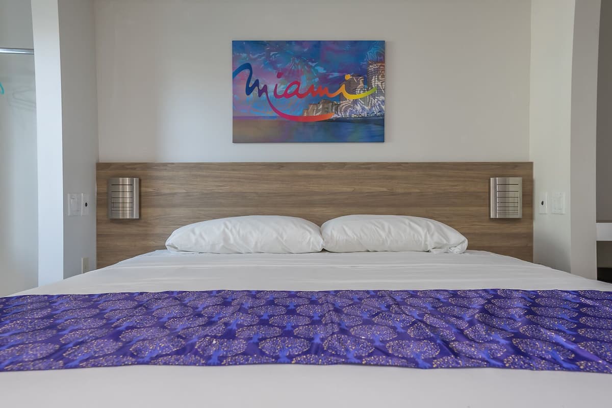 Quarto de hotel com uma cama, almofadas brancas, edredom roxo estampado e uma pintura abstrata colorida na parede.