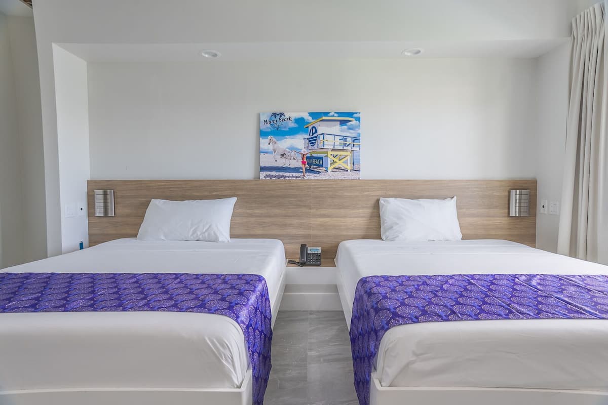 Quarto de hotel com duas camas, uma cabeceira de madeira, um quadro de Miami Beach e um telefone na mesa de cabeceira.