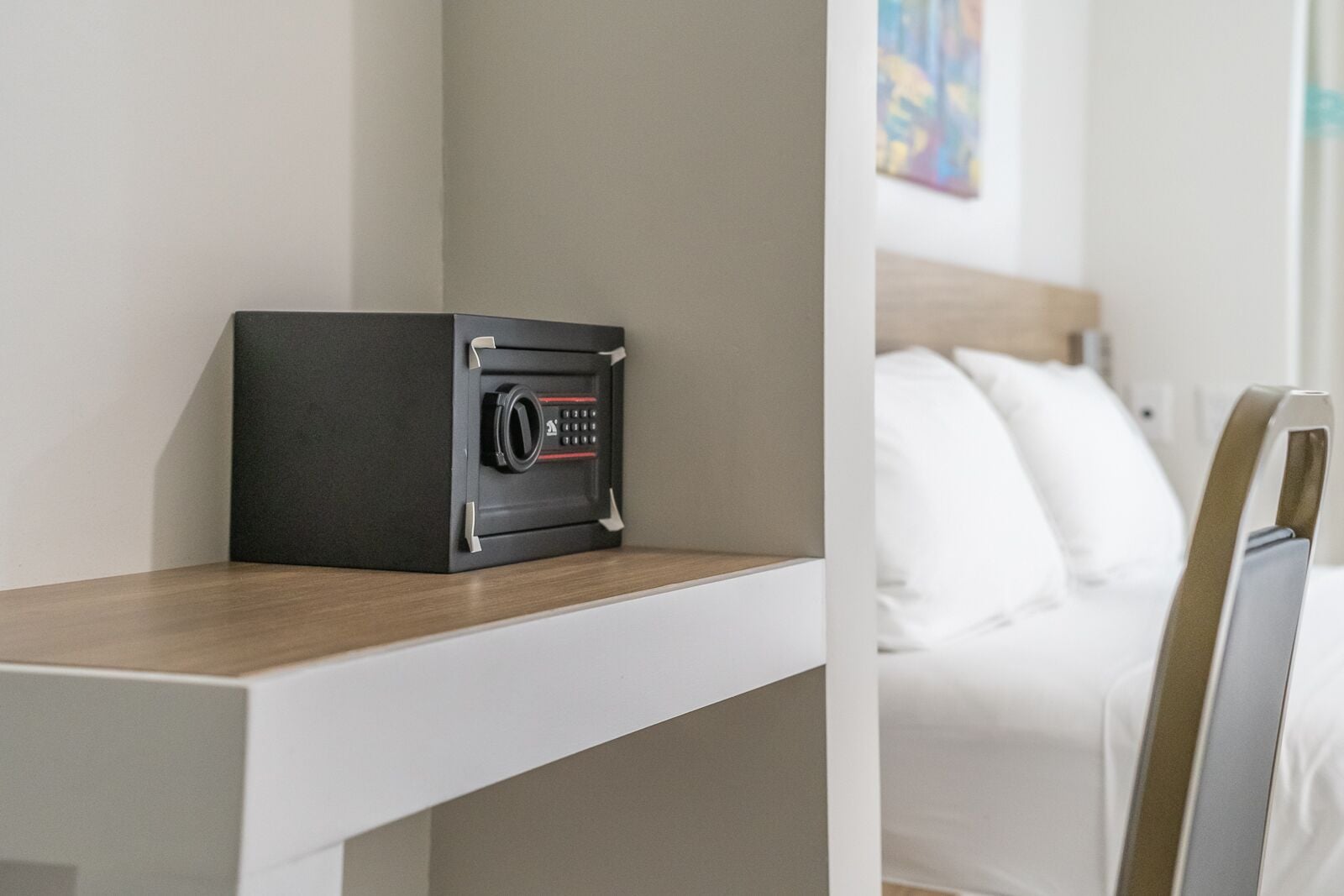 A hotel room with a wooden bedside table, a black safe, and a bed with white bedding.
