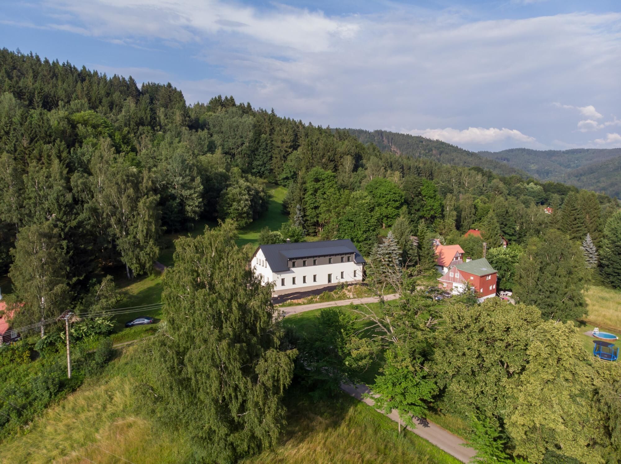 A large white building with a dark roof, surrounded by dense forest and hills.