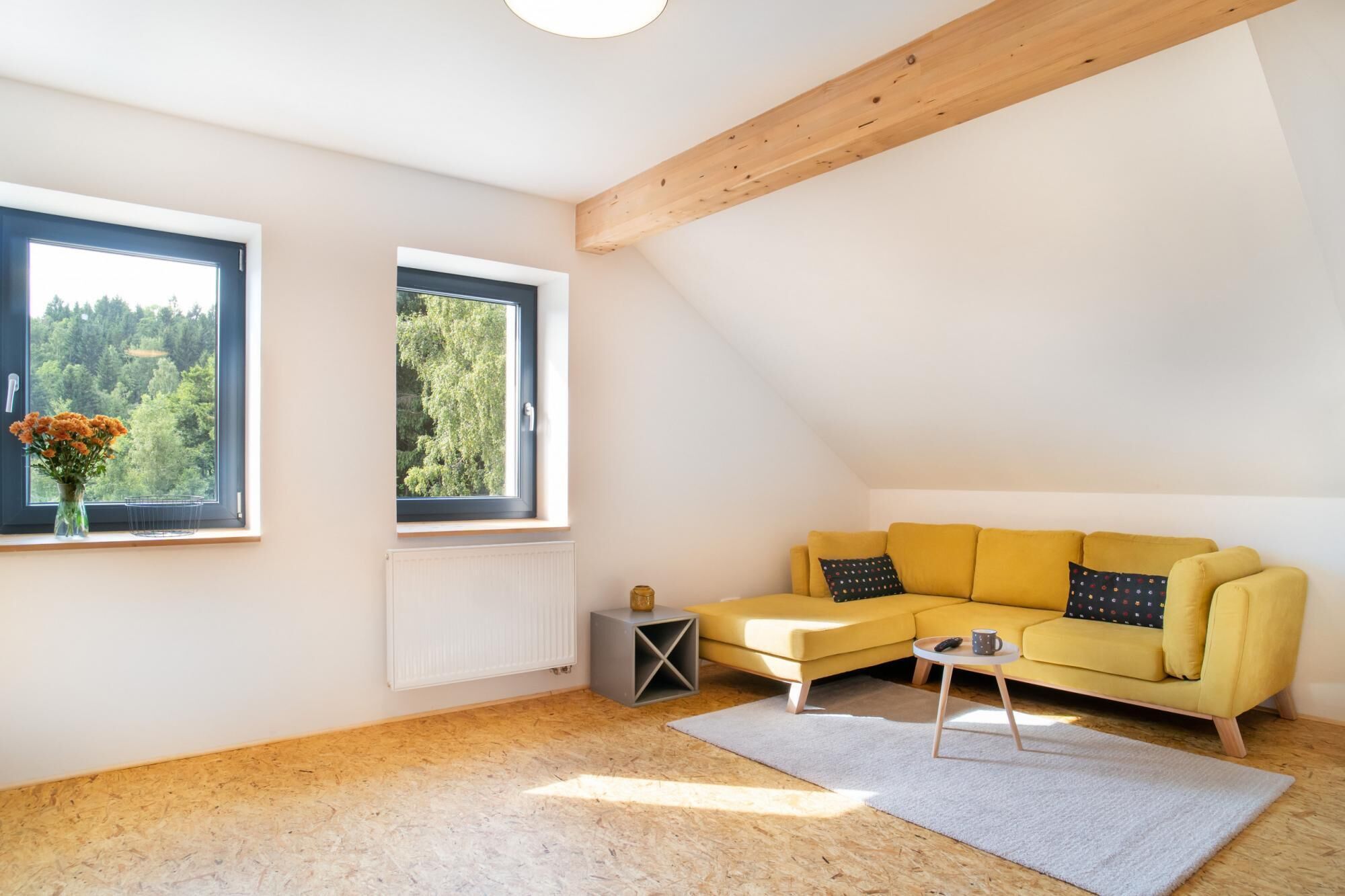Uma sala de estar aconchegante com um sofá amarelo, duas janelas com vista para o verde, um radiador e uma pequena mesa lateral com uma xícara.