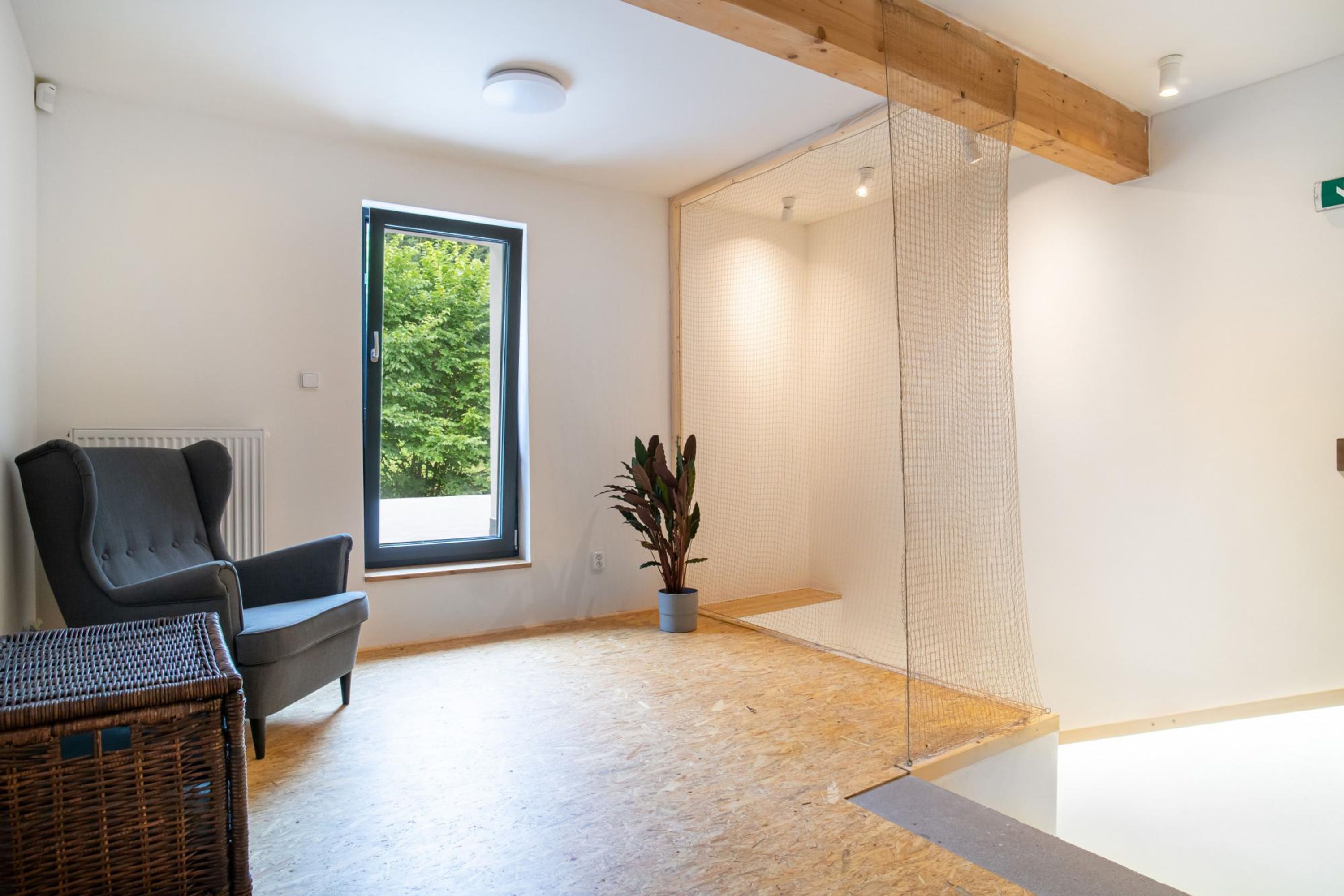 Quarto moderno com uma janela grande, uma poltrona cinza, uma mesinha de vime e uma planta em vaso.