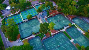 Vue aérienne d’un complexe sportif comprenant plusieurs courts de tennis, entourés d’arbres et de zones de stationnement.