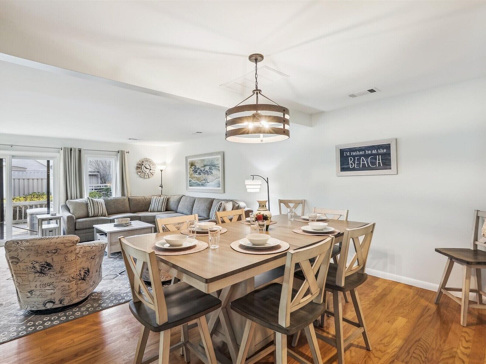 A dining area with a wooden table set for four, surrounded by wooden chairs. A chandelier hangs above the table, and there is a framed sign on the wall that reads "I'd rather be at the beach."