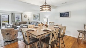 Un coin repas avec une table en bois, dressée pour quatre personnes, entourée de chaises en bois. Un lustre est suspendu au-dessus de la table, et un panneau encadré est accroché au mur, sur lequel on peut lire : « Je préférerais être à la plage. »
