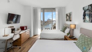 A hotel room with a bed, a desk, a chair, a TV, a window with curtains, and artwork on the wall. - Infinite View| Balcony + Pool + Gym +  Near Beach (Hallandale Beach)