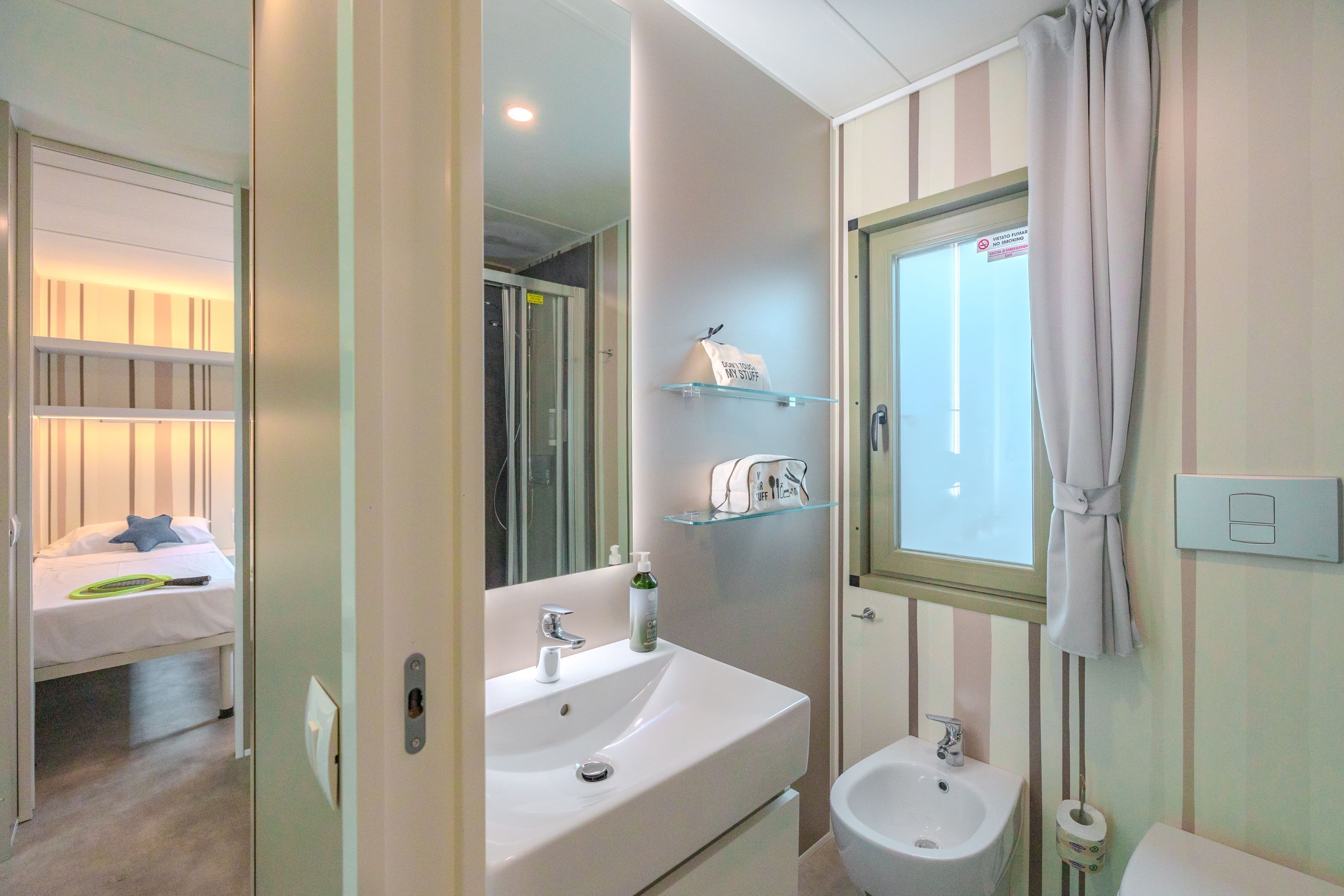 A bathroom with a sink, mirror, and two shelves; a bed with a pillow and a green object; and a window with a curtain.
