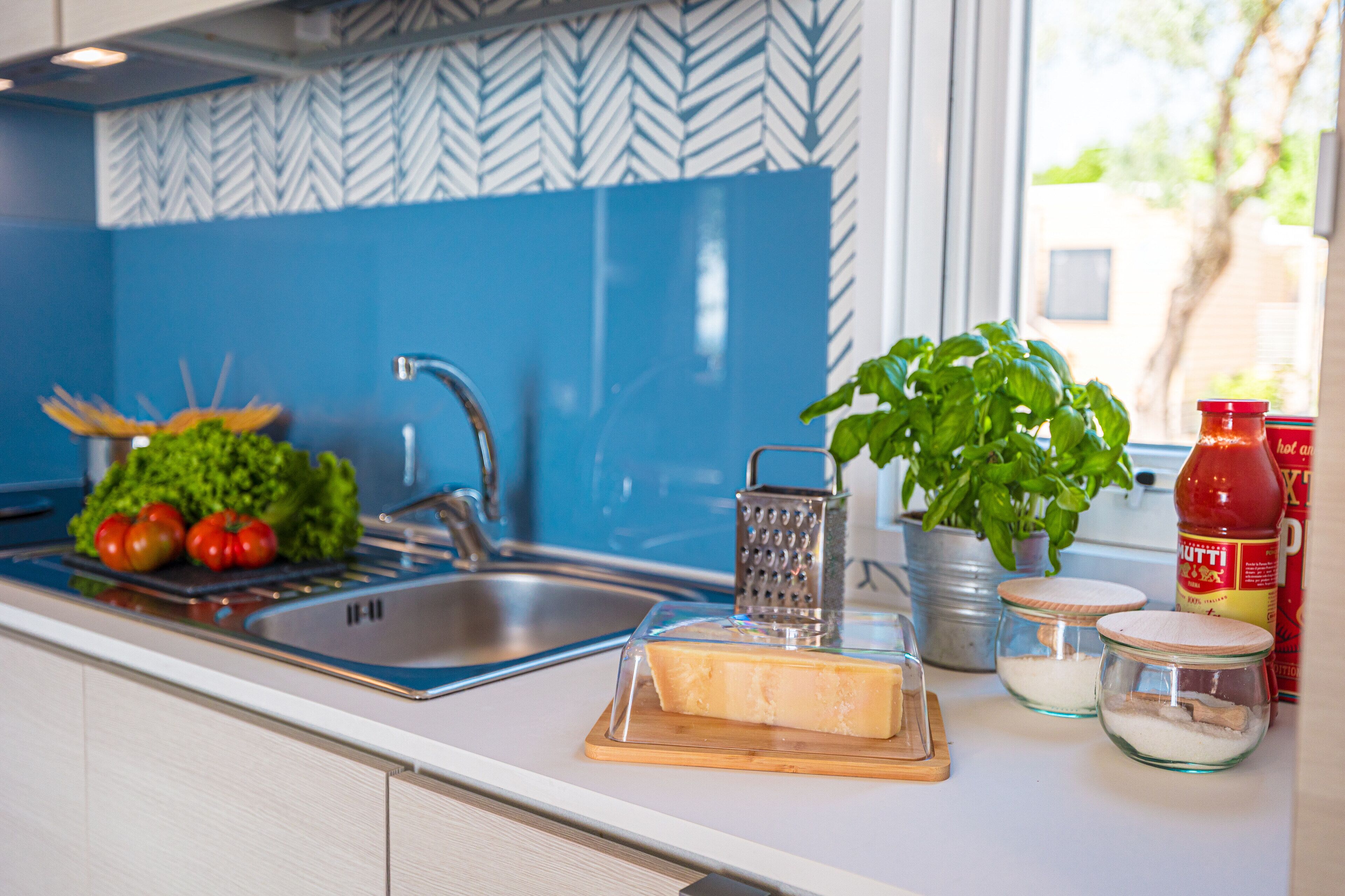 Eine Küche mit blauer Rückwand, einem Spülbecken, einem Schneidebrett mit Tomaten, einer Reibe, einer Pflanze und Gläsern voller Zutaten.