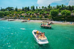 Un bateau avec des personnes à bord, naviguant sur une mer turquoise et limpide, un quai et une plage parsemée de parasols et fréquentée par des gens.