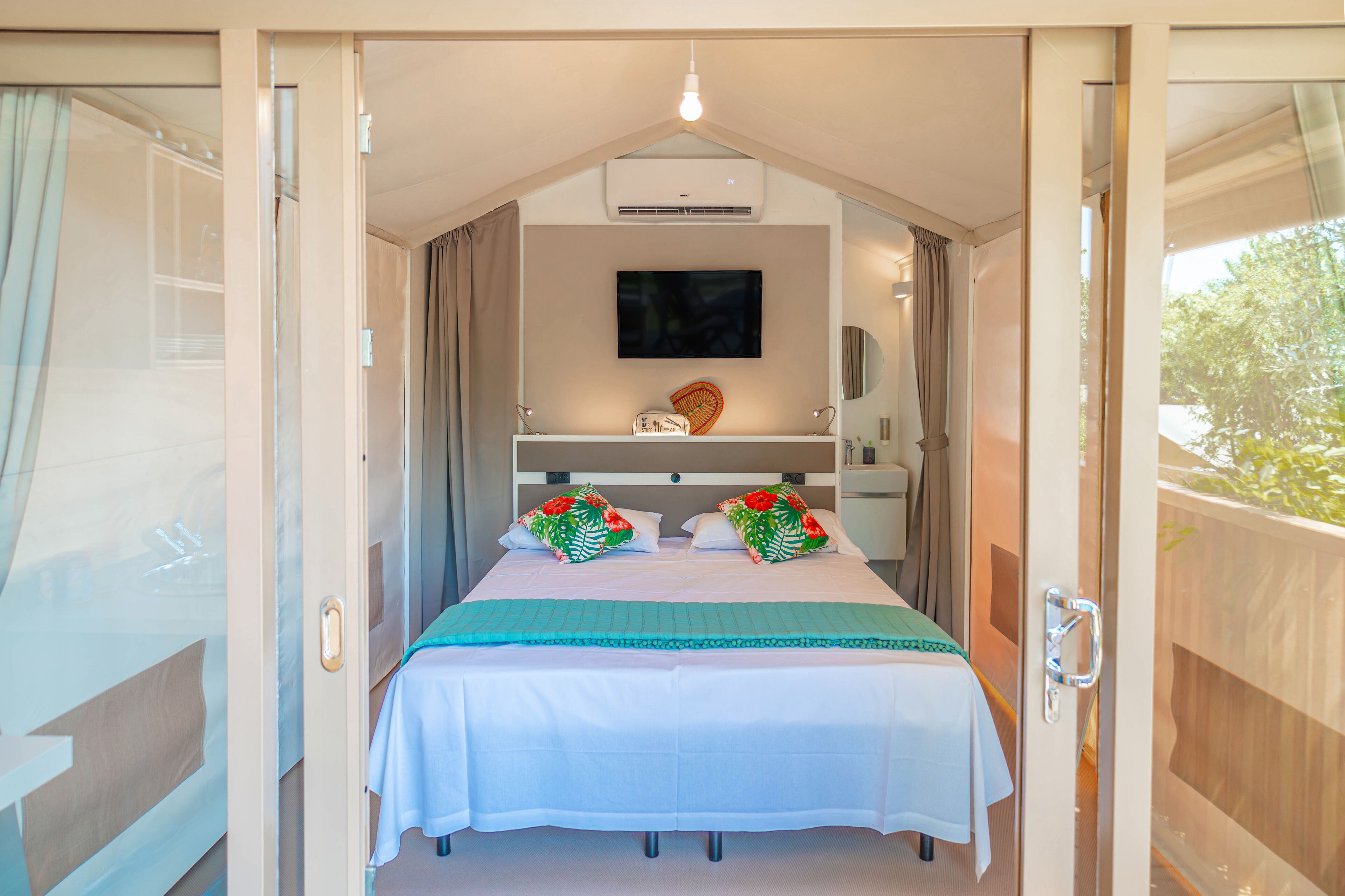 Un dormitorio con cama, televisor de pantalla plana, una mesita con un objeto decorativo y una ventana con cortinas.