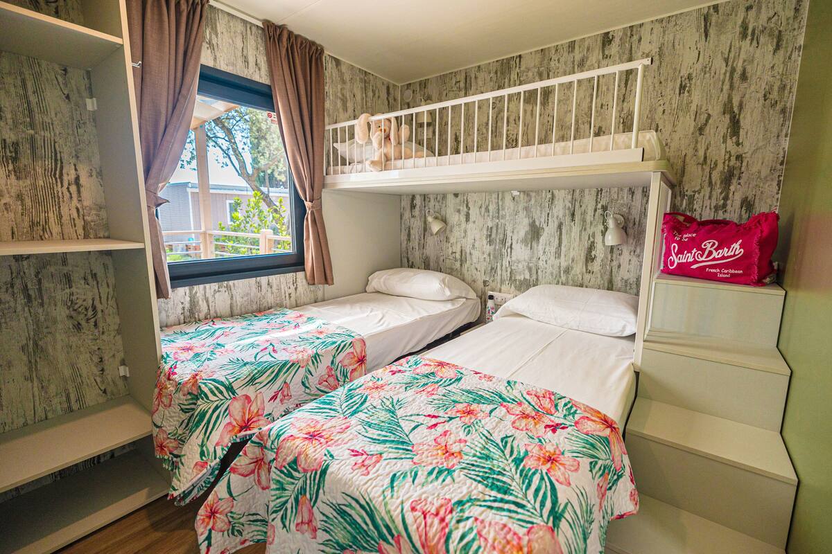 Una habitación con dos literas, ropa de cama floral, una ventana con cortinas y una lámpara de pared.