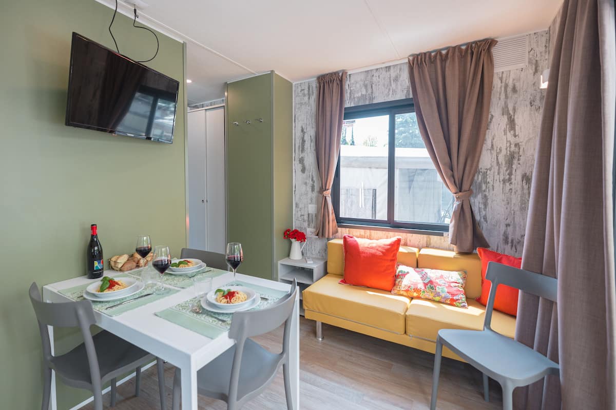 Un comedor con una mesa dispuesta para dos personas, un sofá con cojines, un televisor montado en la pared y una ventana con cortinas.