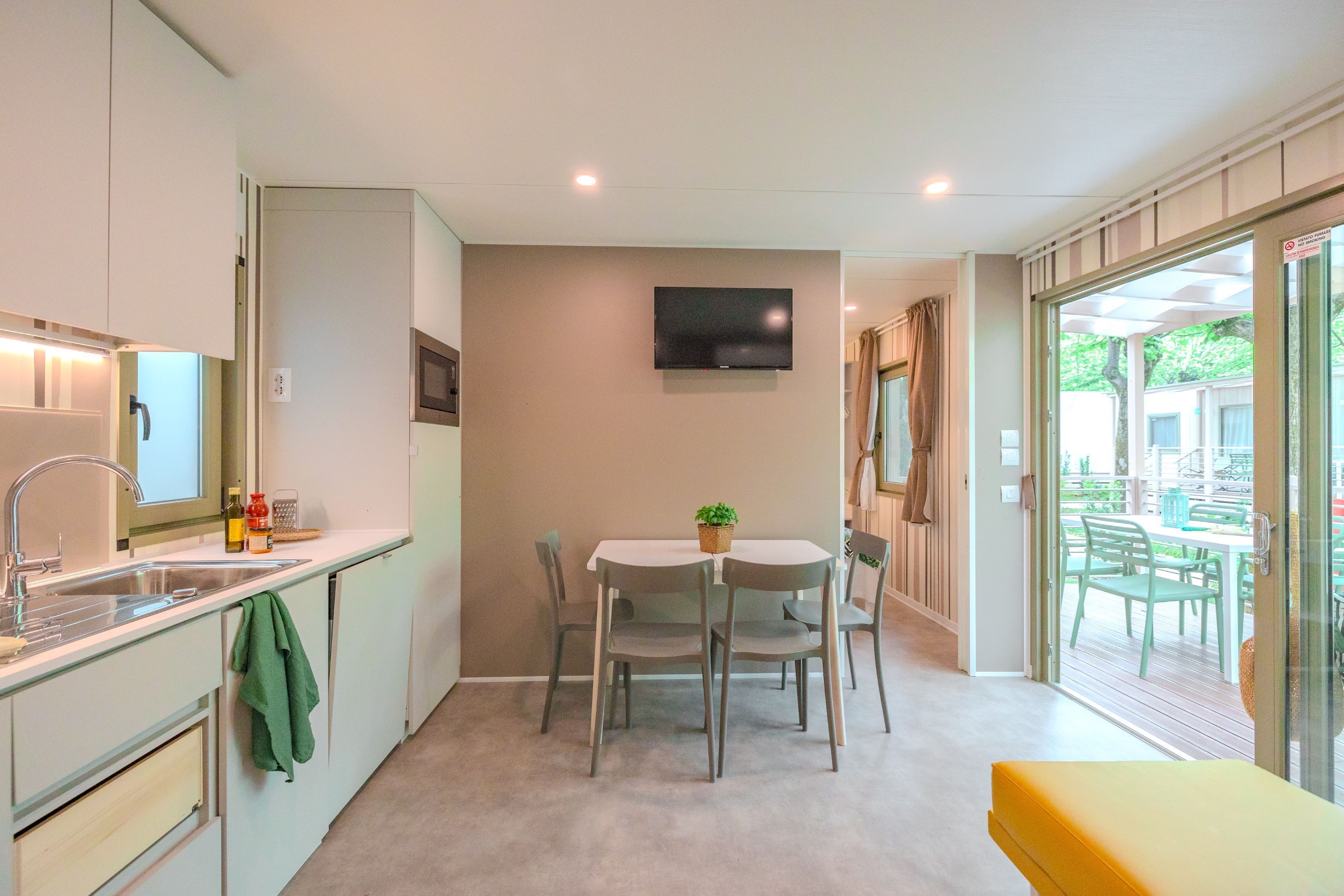 A compact kitchen with a dining area, a flat-screen TV mounted on the wall, and a sliding glass door leading to an outdoor patio.