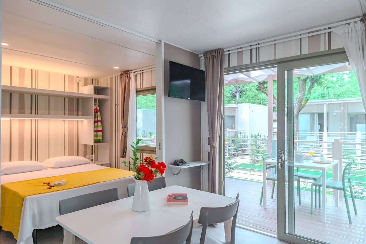 Habitación de hotel con cama, una mesa con un florero, un televisor montado en la pared y un ventanal corredero que da a un espacio exterior con mesa y sillas.