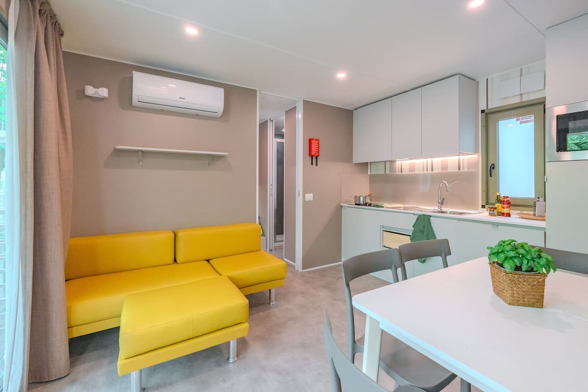 Un espacio habitable compacto con un sofá amarillo, una mesa de comedor, una zona de cocina y una ventana pequeña.