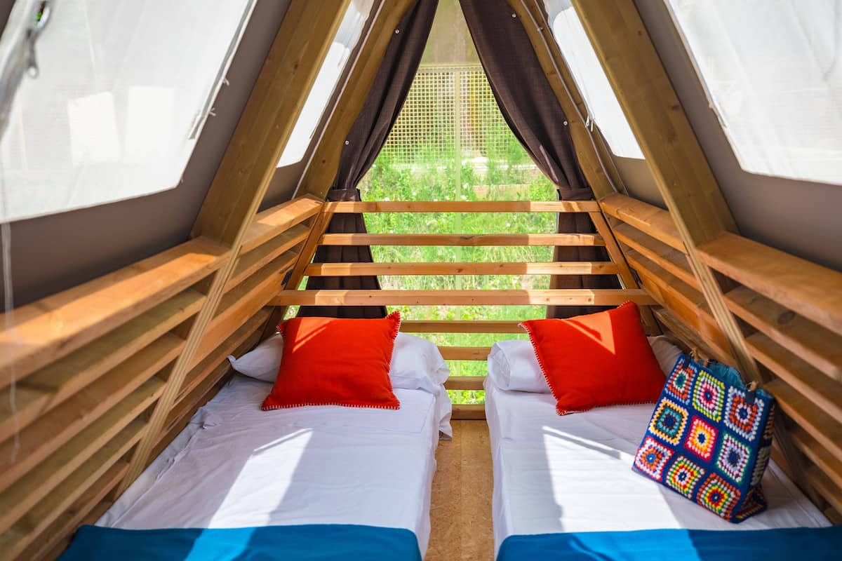 Una habitación tipo tienda de campaña, acogedora y triangular, con dos camas, cada una con ropa de cama blanca y almohadas rojas.