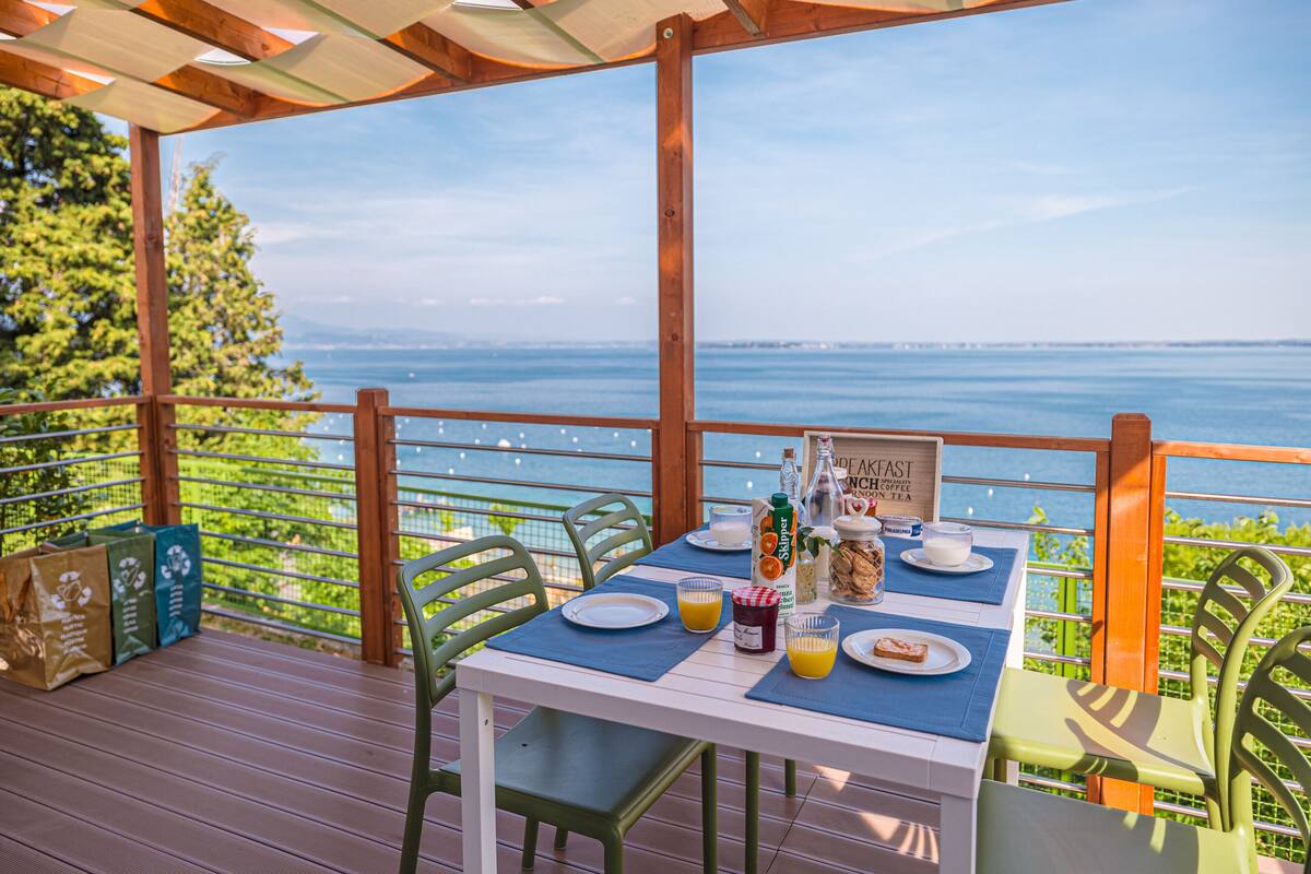 Una mesa lista para el desayuno con vista al mar, una terraza de madera y sillas verdes.