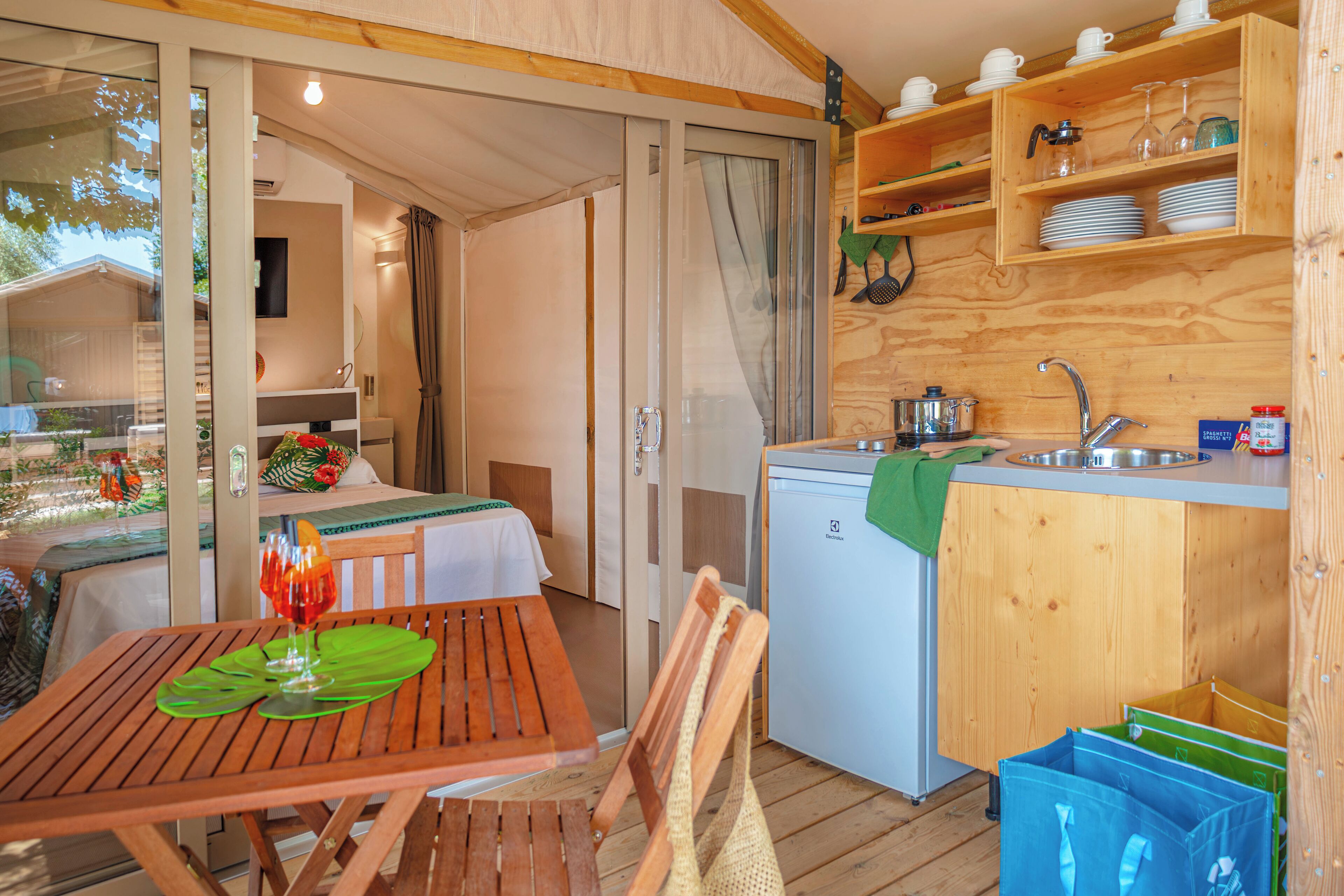A compact kitchen with wooden cabinets, a sink, and a small refrigerator. There is a dining area with a table and chairs, and a view through a glass door to an outdoor area.
