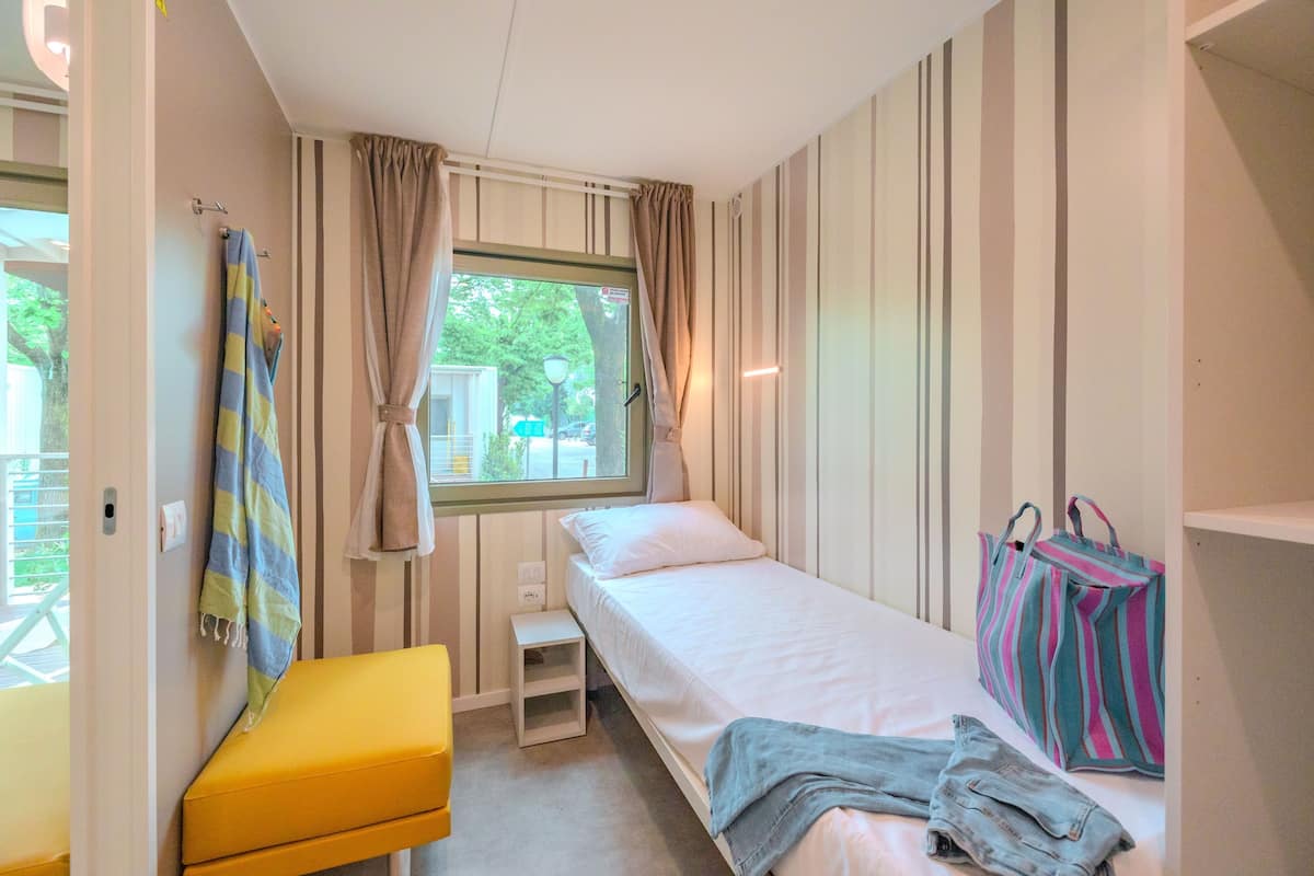 Un dormitorio pequeño y ordenado con una cama individual, una mesita de noche, un banco y una ventana con cortinas.
