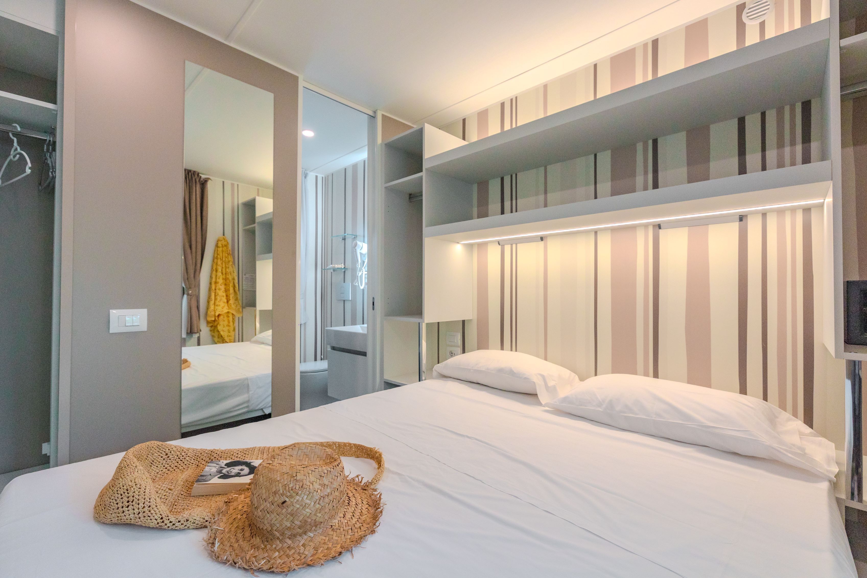 A neatly made bed with a woven basket and a book, a mirror, and a striped wall with built-in shelves.