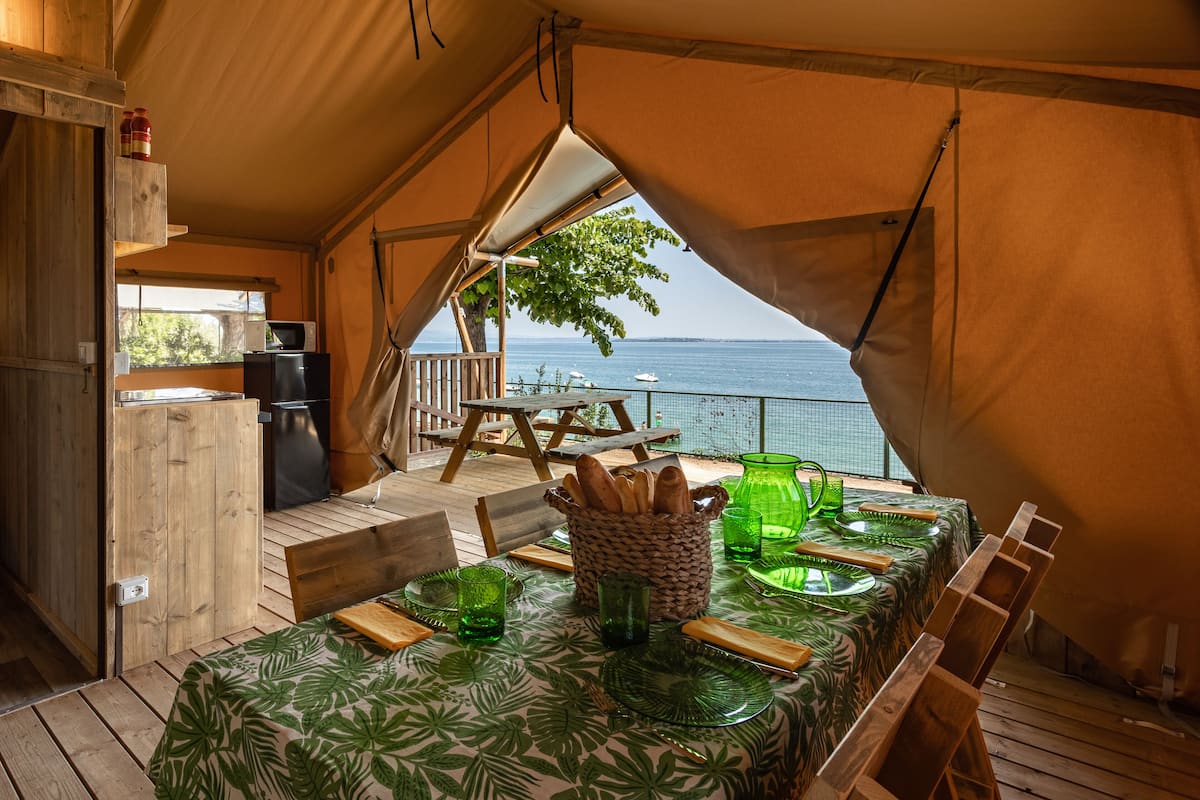 Un comedor cubierto con carpa, una mesa preparada para dos, una cesta de pan y vista al mar.
