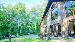 En modern stuga i trä med stora fönster, en uteplats med utemöbler och en grill, belägen i ett skogsområde.