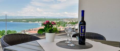 A balcony with a table, two wine glasses, a bottle of wine, a potted plant, and a view of the sea and buildings.