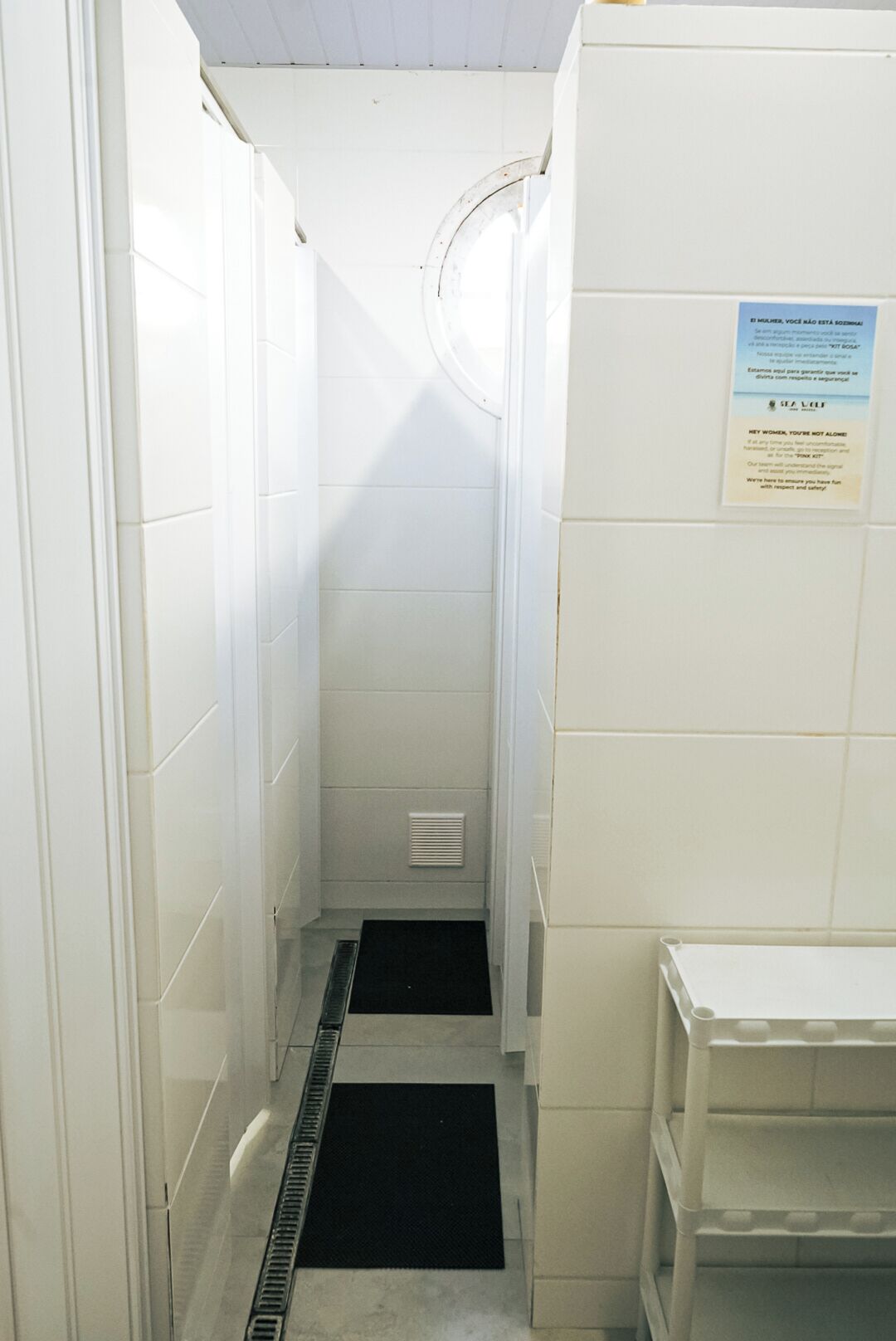 A narrow, tiled hallway with a vent and a black mat at the end. A sign on the wall reads 'HEY WOMEN, YOU'RE NOT ALONE.'