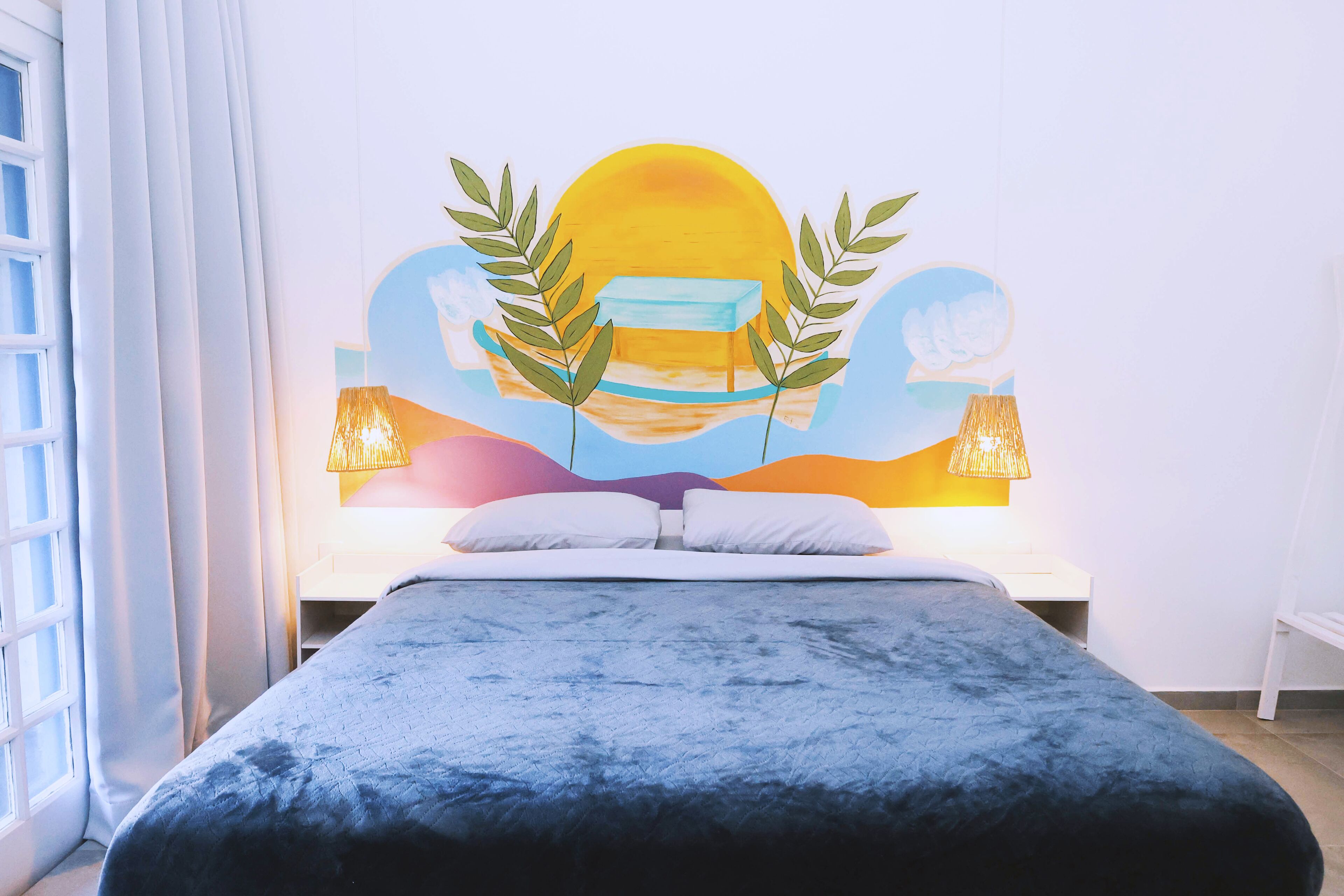 Una cama con cubierta azul, un cuadro de un paisaje con sol y árboles, y dos lámparas a cada lado de la cama.