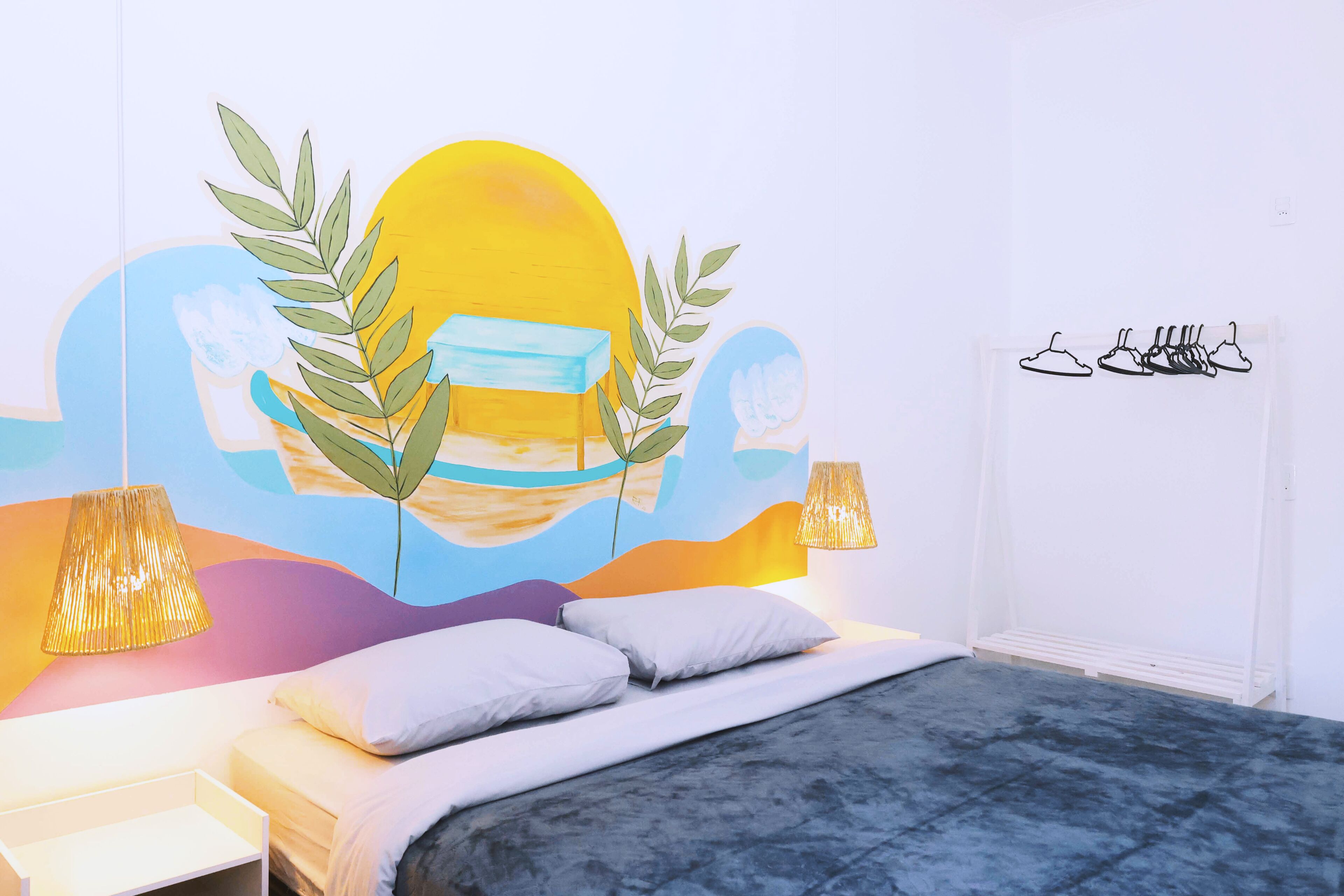 A bedroom with a bed, bedside lamps, a wall art of a sun and leaves, and a wall-mounted shoe rack.
