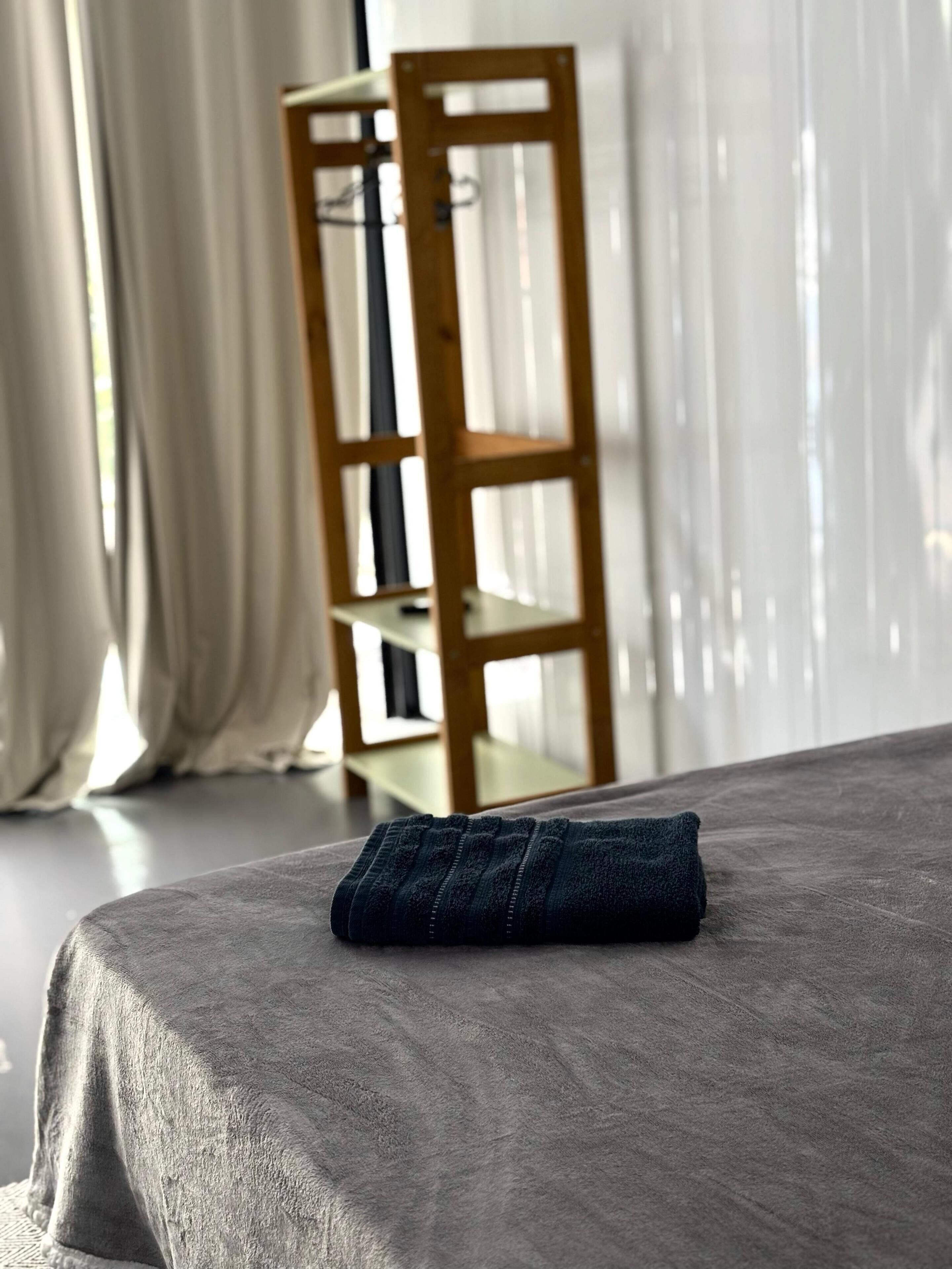A neatly made bed with a grey bedspread and a folded dark blue towel on top. In the background, there is a wooden chair with a shelf and sheer curtains.